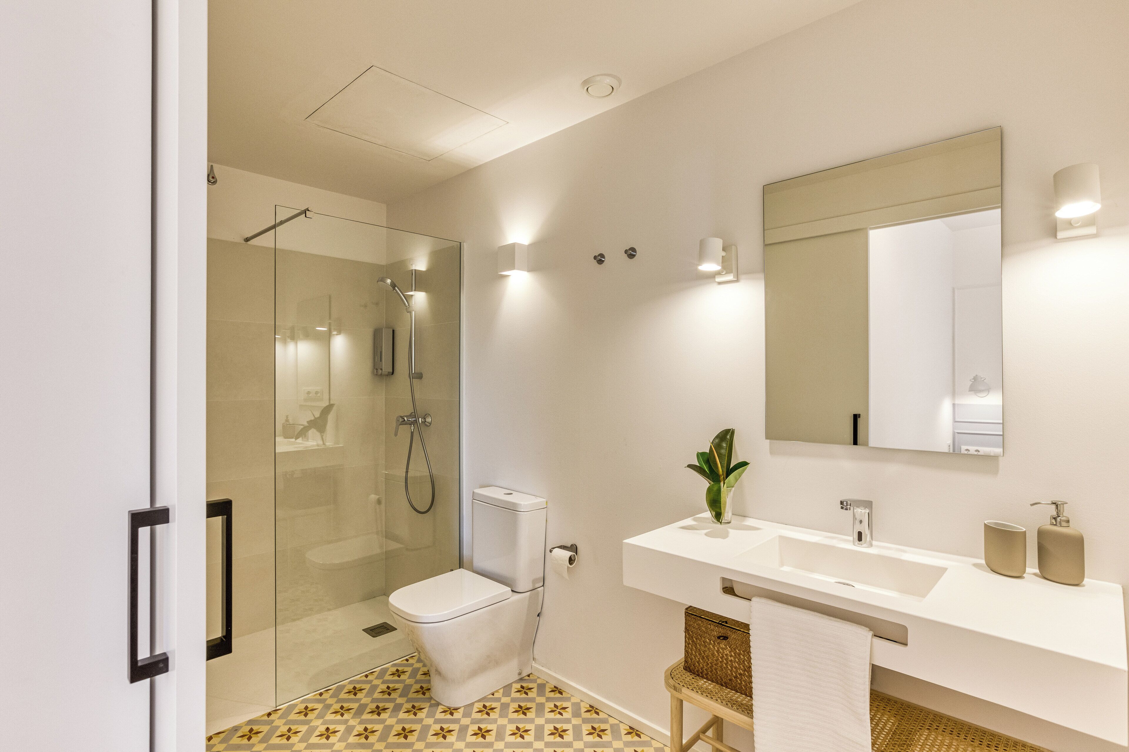 A modern bathroom with a glass-enclosed shower, a white toilet, a sink with a mirror, and a patterned floor.