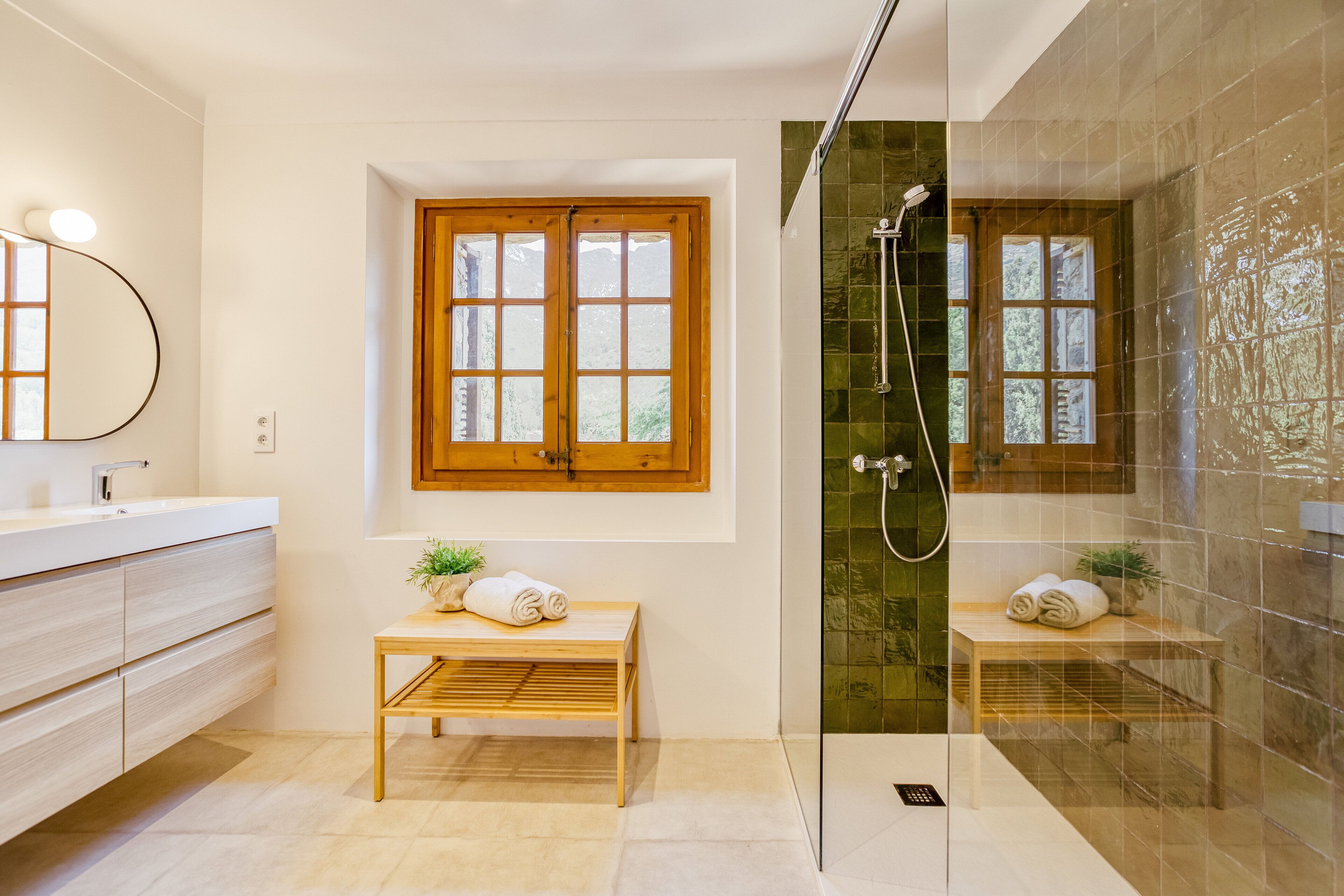 Un baño moderno con tocador de madera, un banco, un espejo grande, una ventana y zona de ducha con paredes de vidrio.