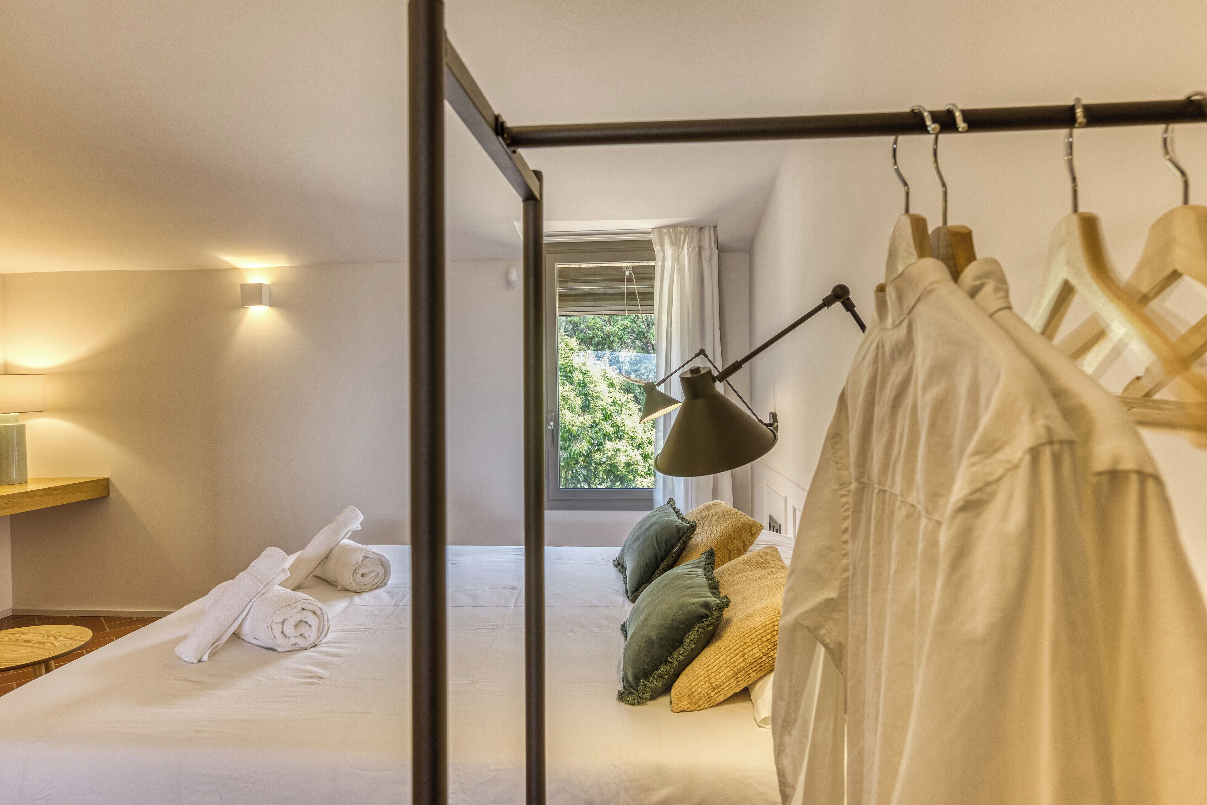 Un dormitorio con cama, almohadas, un perchero con ropa, una lámpara y una ventana con cortinas.
