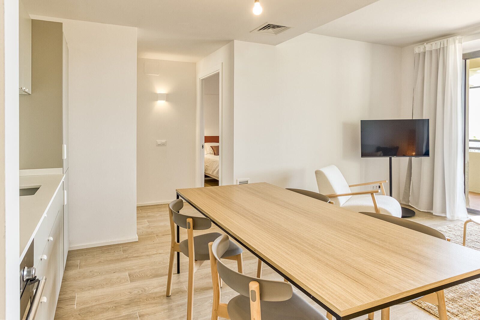 Una sala de estar moderna con una mesa de comedor de madera, dos sillas, un televisor de pantalla plana y vista a un dormitorio por una puerta abierta.