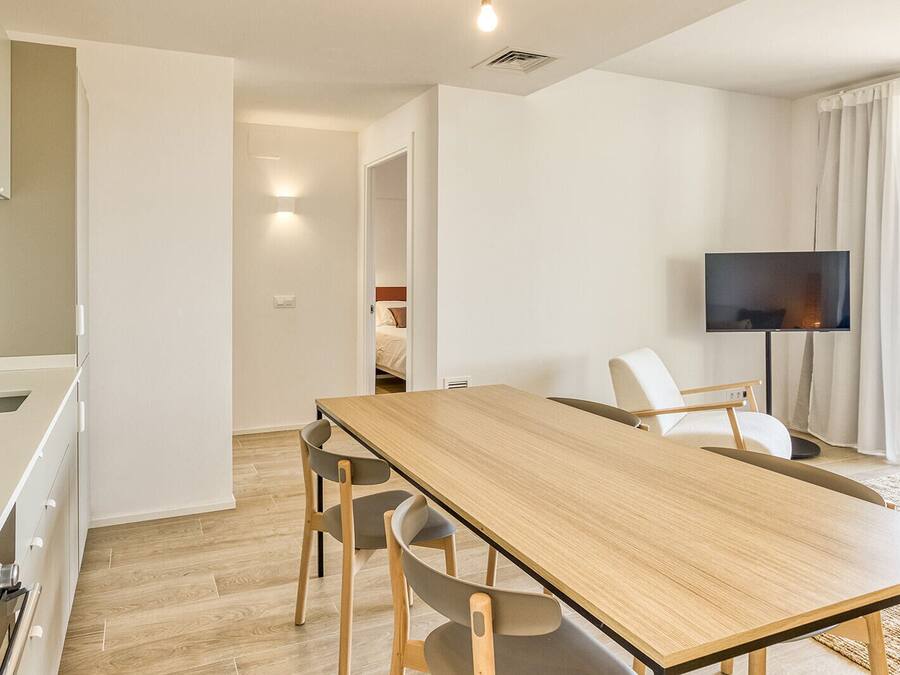 Una sala de estar moderna con una mesa de comedor de madera, dos sillas, un televisor de pantalla plana y vista a un dormitorio por una puerta abierta.