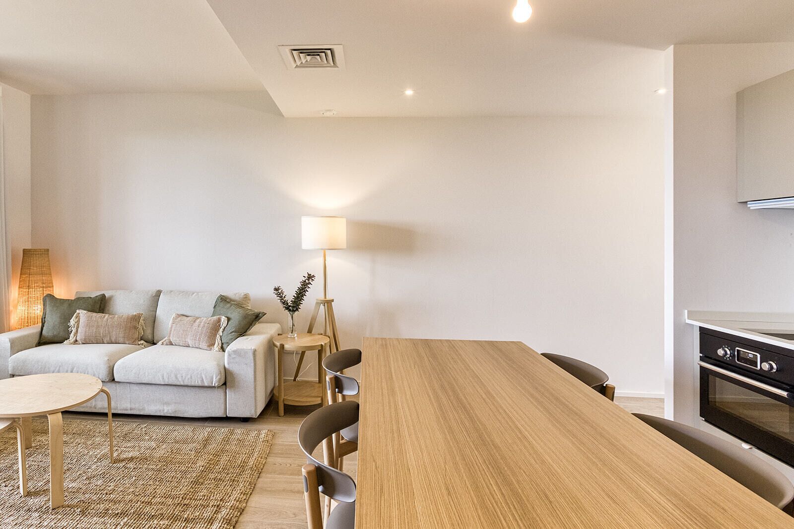 Una sala moderna con un sofá, una mesa redonda de centro y una mesa de comedor con sillas. Se observa una lámpara, una lámpara de pie y una zona de cocina con horno.