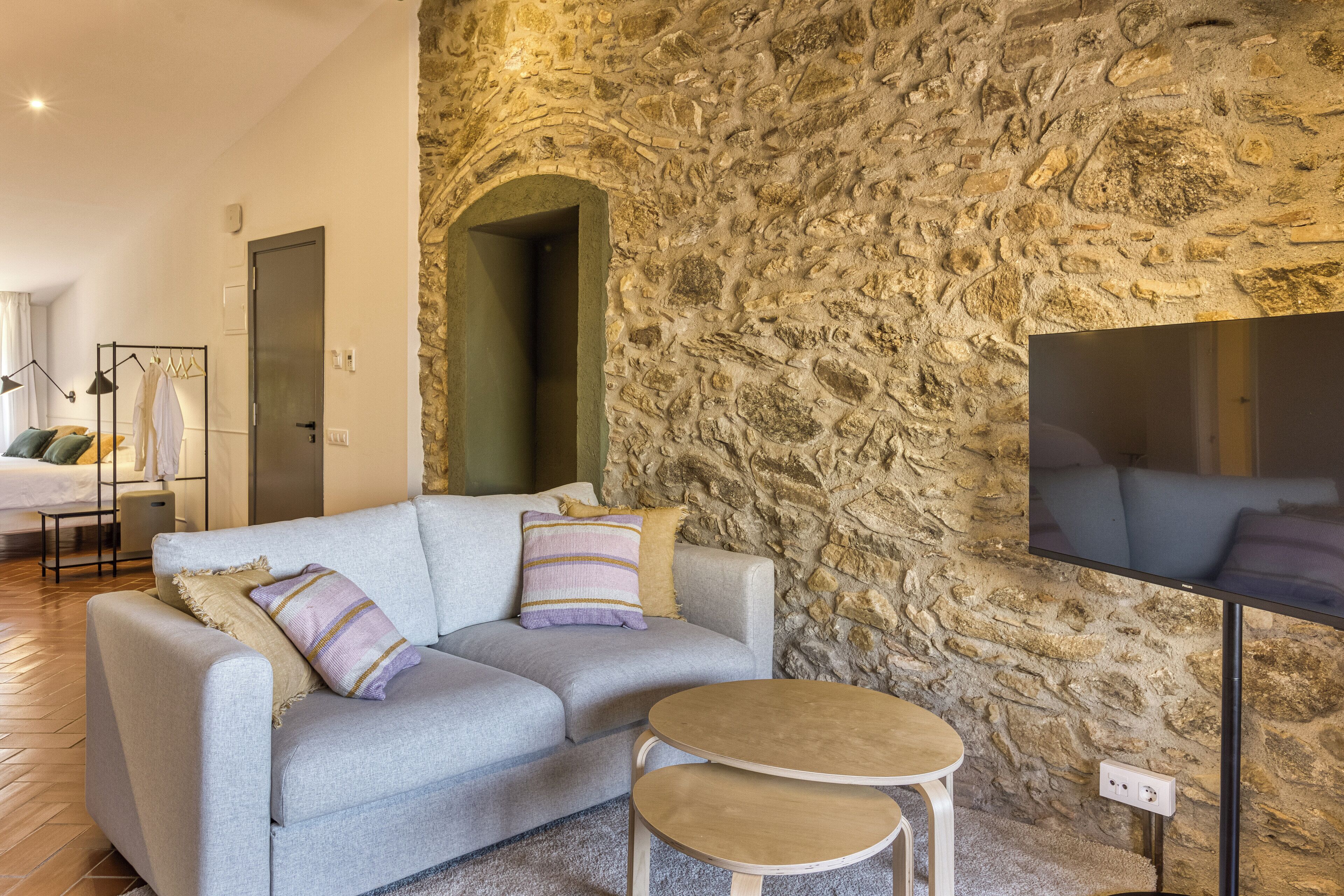 Una sala de estar moderna con una pared de piedra, un sofá gris con cojines a rayas, una mesita redonda y un televisor de pantalla plana montado en la pared.