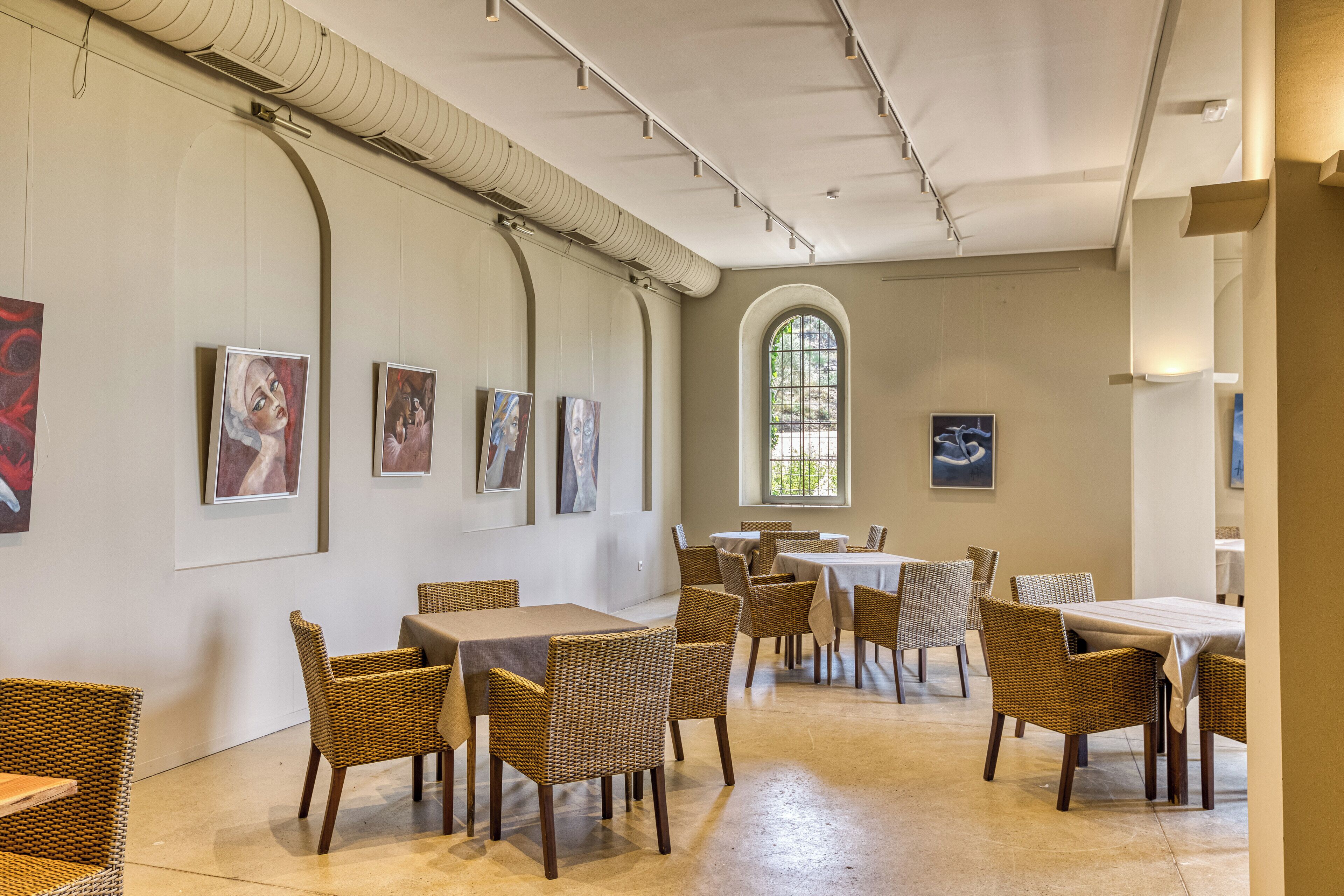 Un área de comedor con sillas y mesas de mimbre, obras de arte en las paredes y un amplio ventanal en arco.