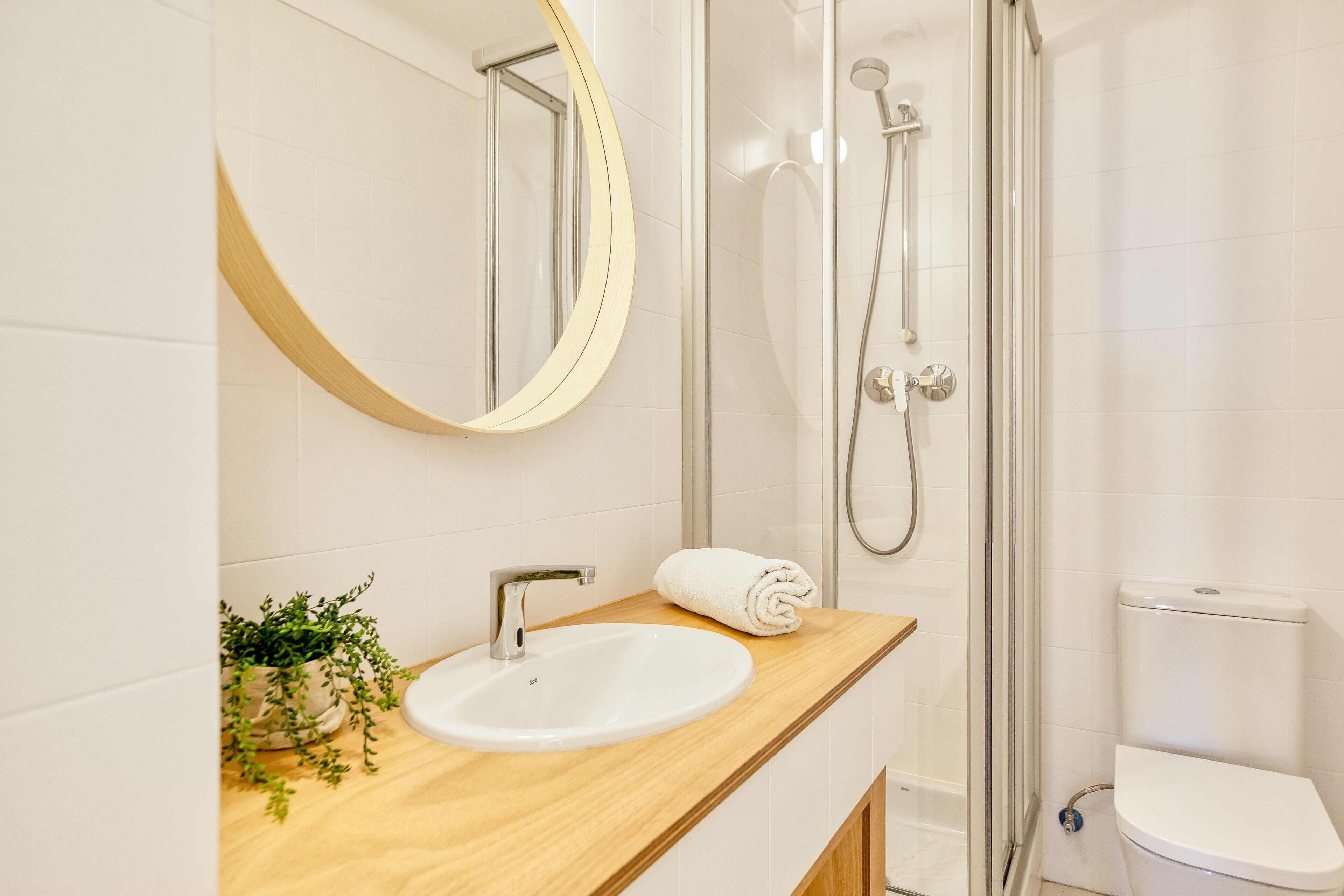A bathroom with a wooden vanity, a white sink, a round mirror, a glass shower enclosure, and a toilet.