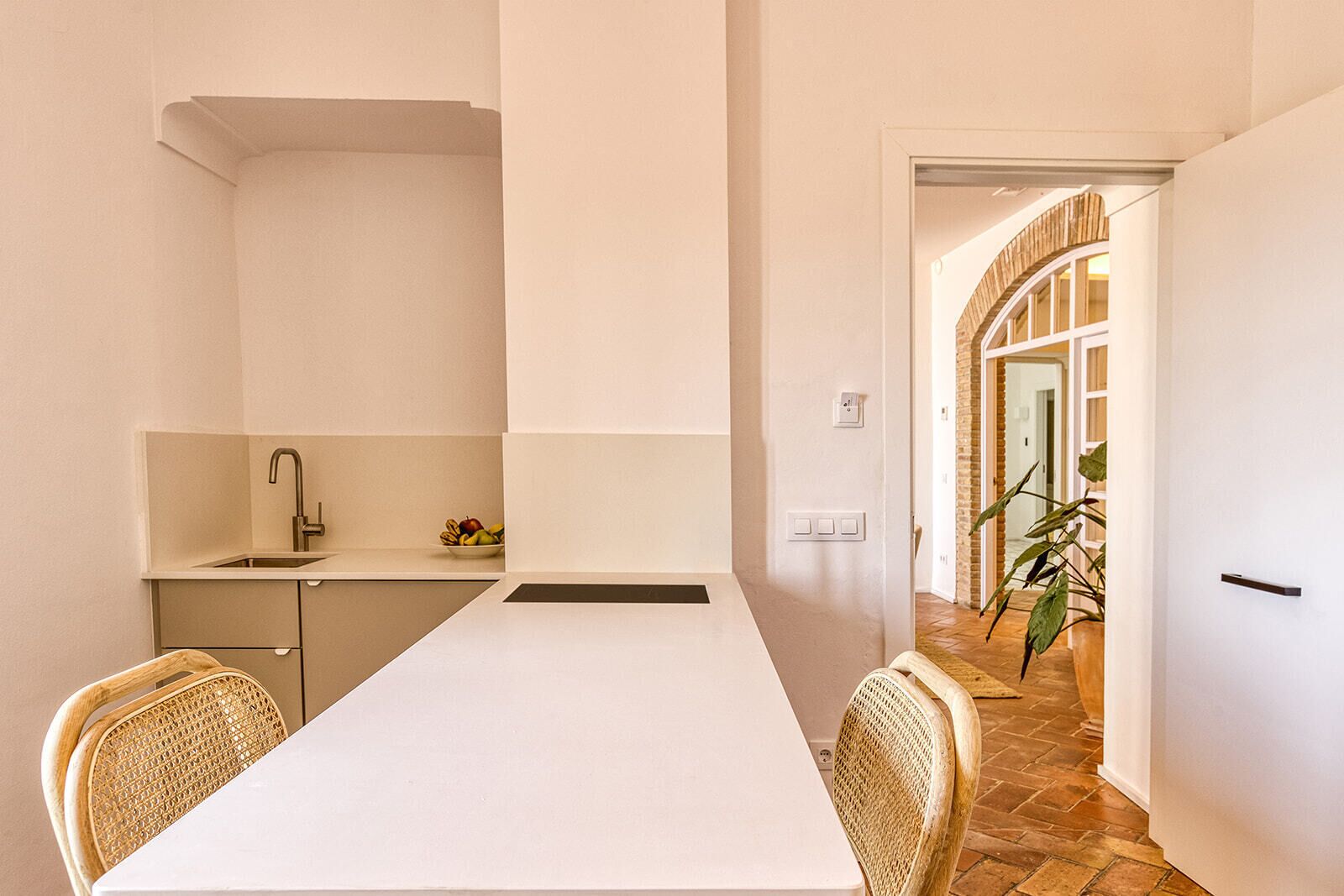 Une cuisine moderne avec un plan de travail blanc, un évier et un saladier rempli de fruits. On y trouve des chaises en osier et une porte qui donne sur un couloir où se trouve une plante.