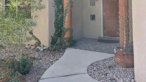 L’entrée d’un immeuble est marquée par un chemin en béton menant à une porte en bois, le tout étant entouré d’un petit jardin agrémenté d’un arbre et de plantes en pot.
