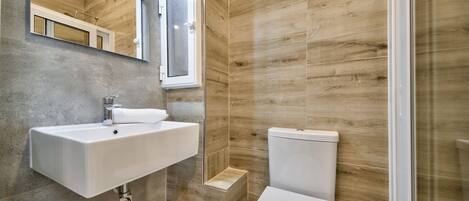 Un baño moderno con lavamanos, espejo y retrete blancos, paneles de pared de madera y un pequeño escalón que conduce a la zona de la ducha.