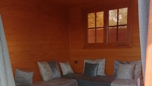 A cozy cabin interior with a wooden ceiling, a sofa set with cushions, a small round table, and a window with a view of trees.