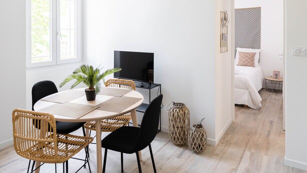Ett modernt och väl upplyst rum med en matplats, en TV och en säng med ett mönstrat sänggavel.