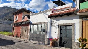 A two-story building with a white facade, a red roof, and a stone wall. There are potted plants and a wooden door.