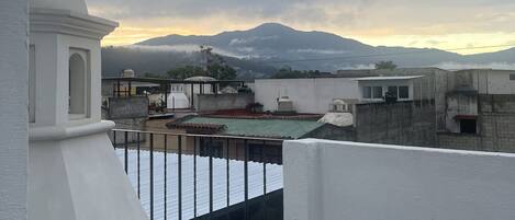Vista desde la azotea con un edificio blanco, barandilla de metal y una cadena montañosa distante bajo cielo nublado.