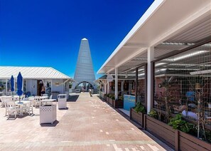 A patio area with outdoor seating, a tall structure in the background, and a covered walkway with plants.