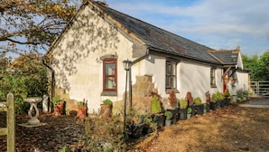 Une maison traditionnelle en pierre, dotée d’un toit en pente, de deux fenêtres aux cadres rouges et d’un jardin agrémenté de plantes en pot et d’une fontaine en pierre.