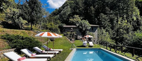 A swimming pool with lounge chairs and umbrellas, surrounded by lush greenery and stone buildings.