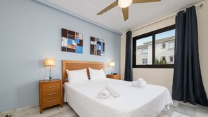 Une chambre d’hôtel avec un grand lit, deux tables de chevet avec des lampes, un ventilateur de plafond, des œuvres d’art au mur et une fenêtre avec des rideaux.