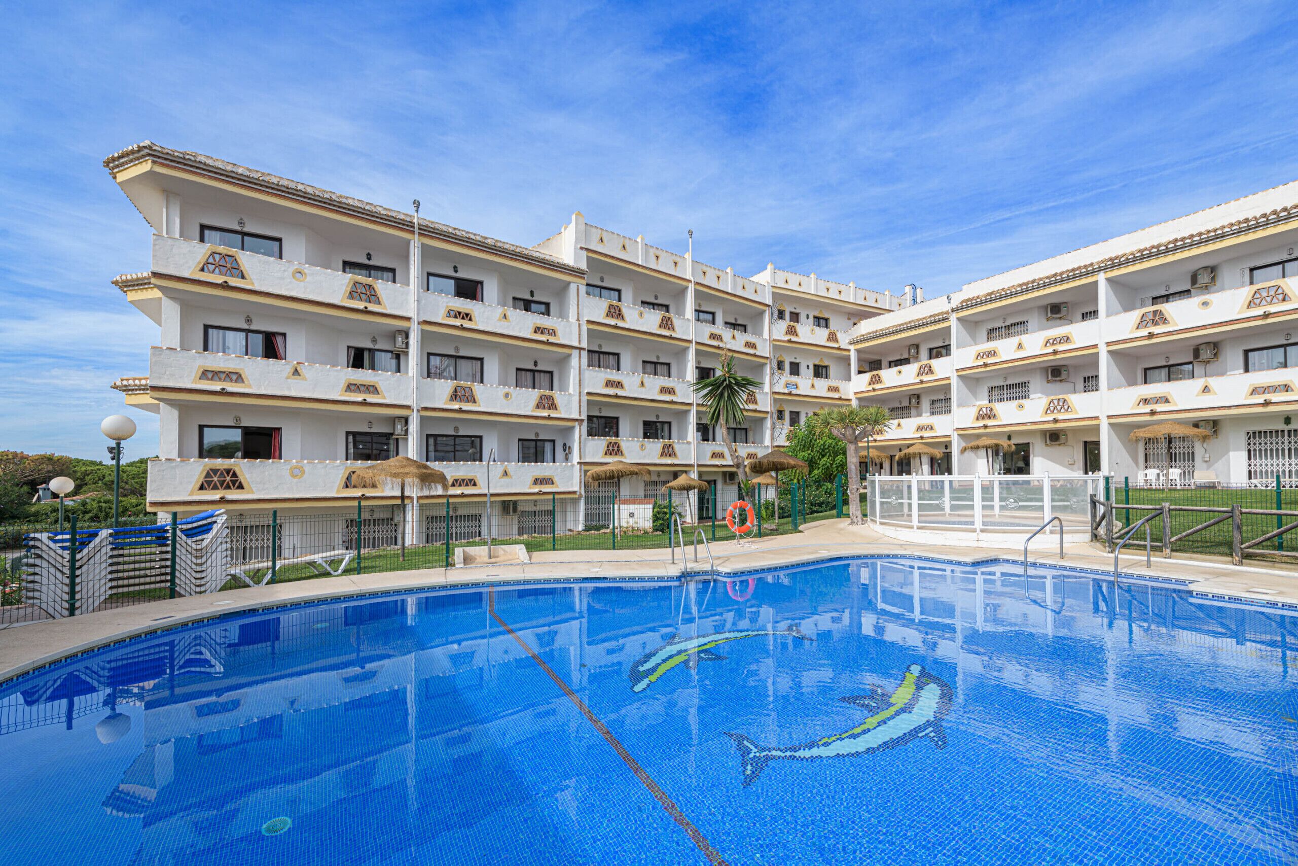 A hotel with a swimming pool, multiple balconies, and a clear blue sky.