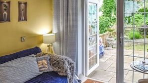 A cozy room with a blue sofa, patterned cushions, a lamp, and a view of a garden through a glass door.