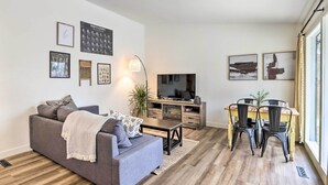 Un salon moderne avec un canapé gris, un plancher en bois, un coin repas avec une table jaune et des chaises noires, et un téléviseur posé sur un meuble en bois.