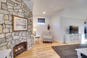 Ein Wohnzimmer mit Kamin aus Stein, einem gerahmten Bild, einem gemusterten Sessel, einem kleinen Tisch mit Lampe und einem Fernseher auf einer weißen Kommode.