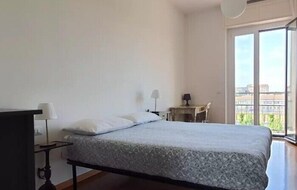 A bedroom with a bed, a nightstand, a lamp, and a view of the city through large windows.
