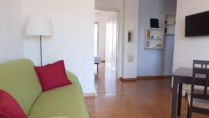 A living room with a green sofa, a red pillow, a white lamp, a dining table with chairs, and a TV mounted on the wall.