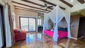 Una habitación amplia con una cama grande bajo dosel, un sofá rojo y un ventanal corredera que da a un balcón.