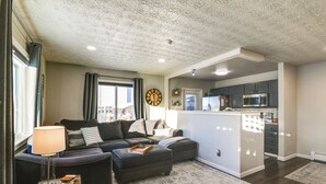A modern living room with a sofa, ottoman, and lamp, leading to a kitchen area with cabinets and appliances. - Updated Townhome: Near Airport & Shopping! (Anchorage)