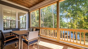 Une terrasse en bois avec une table et des chaises, de grandes fenêtres et une vue sur une forêt et un lac.