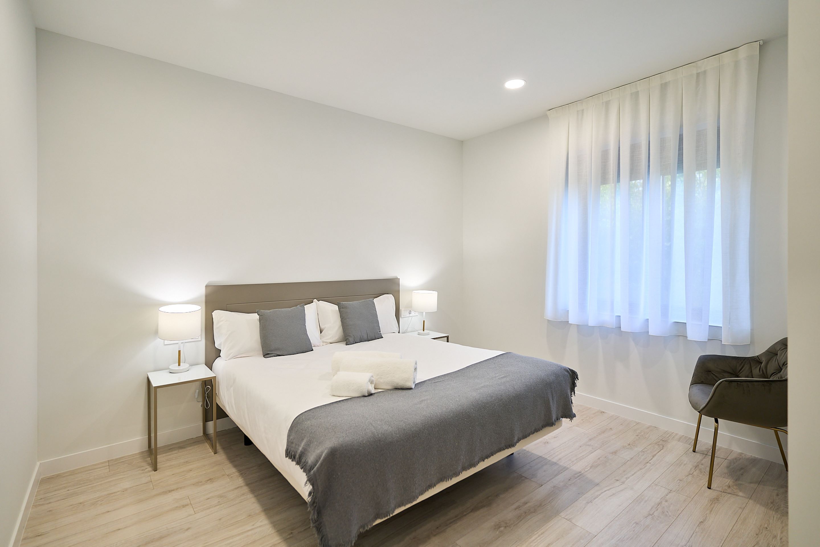 Un dormitorio moderno con una cama grande, lámparas de noche, una silla y un ventanal con cortinas de gasa.
