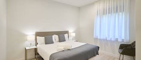 Un dormitorio moderno con una cama grande, lámparas de noche, una silla y un ventanal con cortinas de gasa.