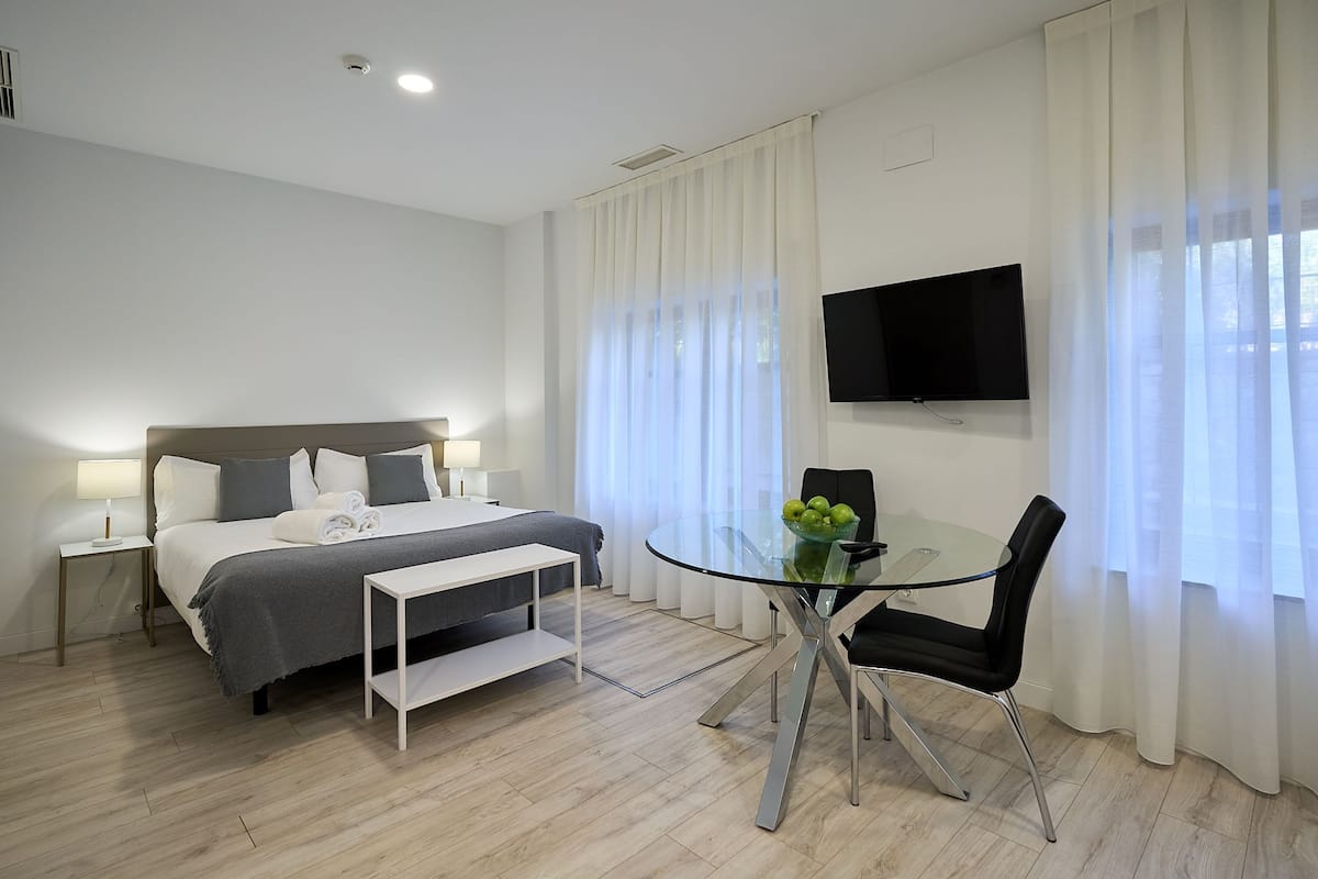 A hotel room with a bed, bedside table, lamp, TV, glass table with chairs, and a window with curtains.