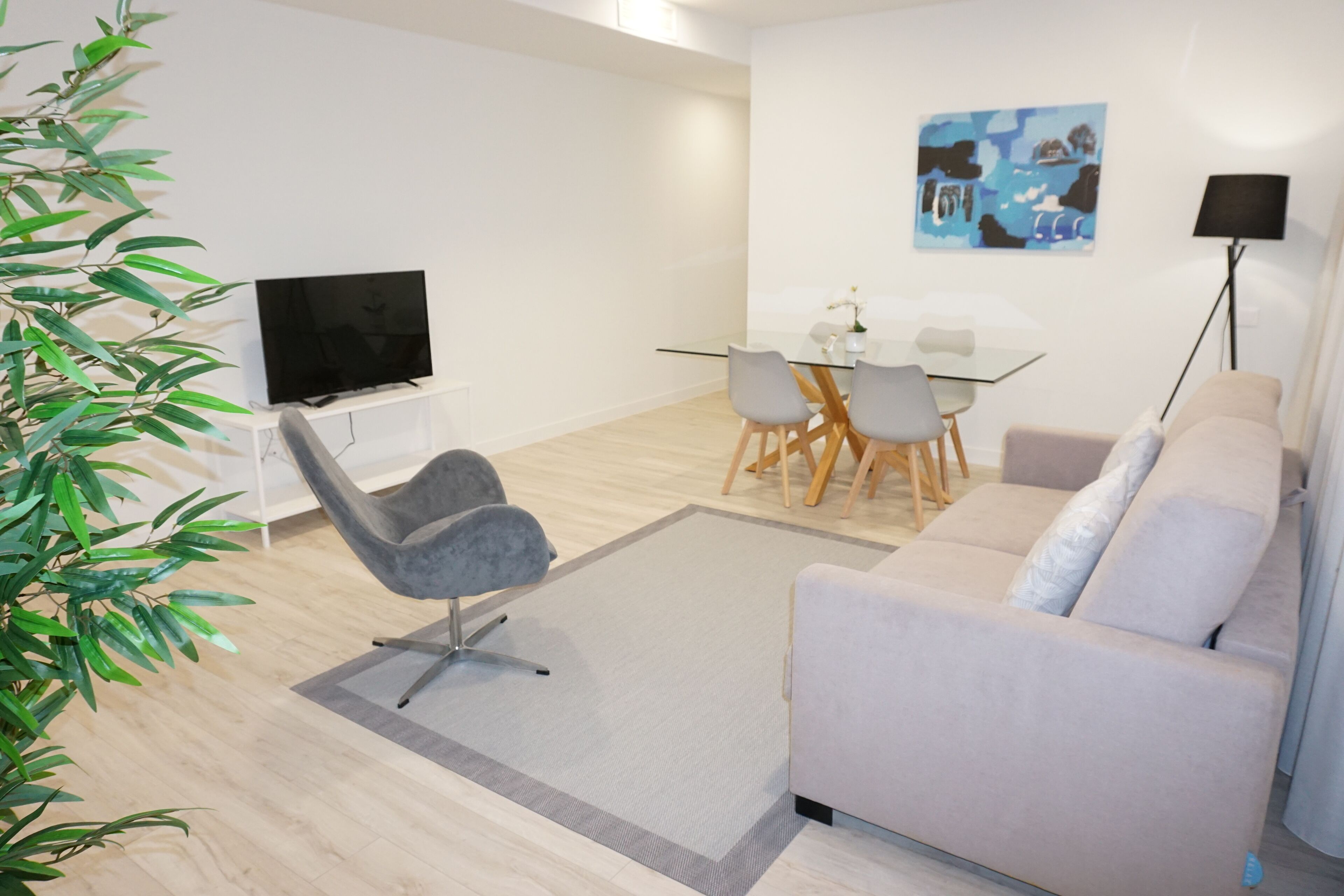 Una sala moderna con un sofá, un televisor, una mesa de vidrio para comer con sillas y una lámpara de pie.