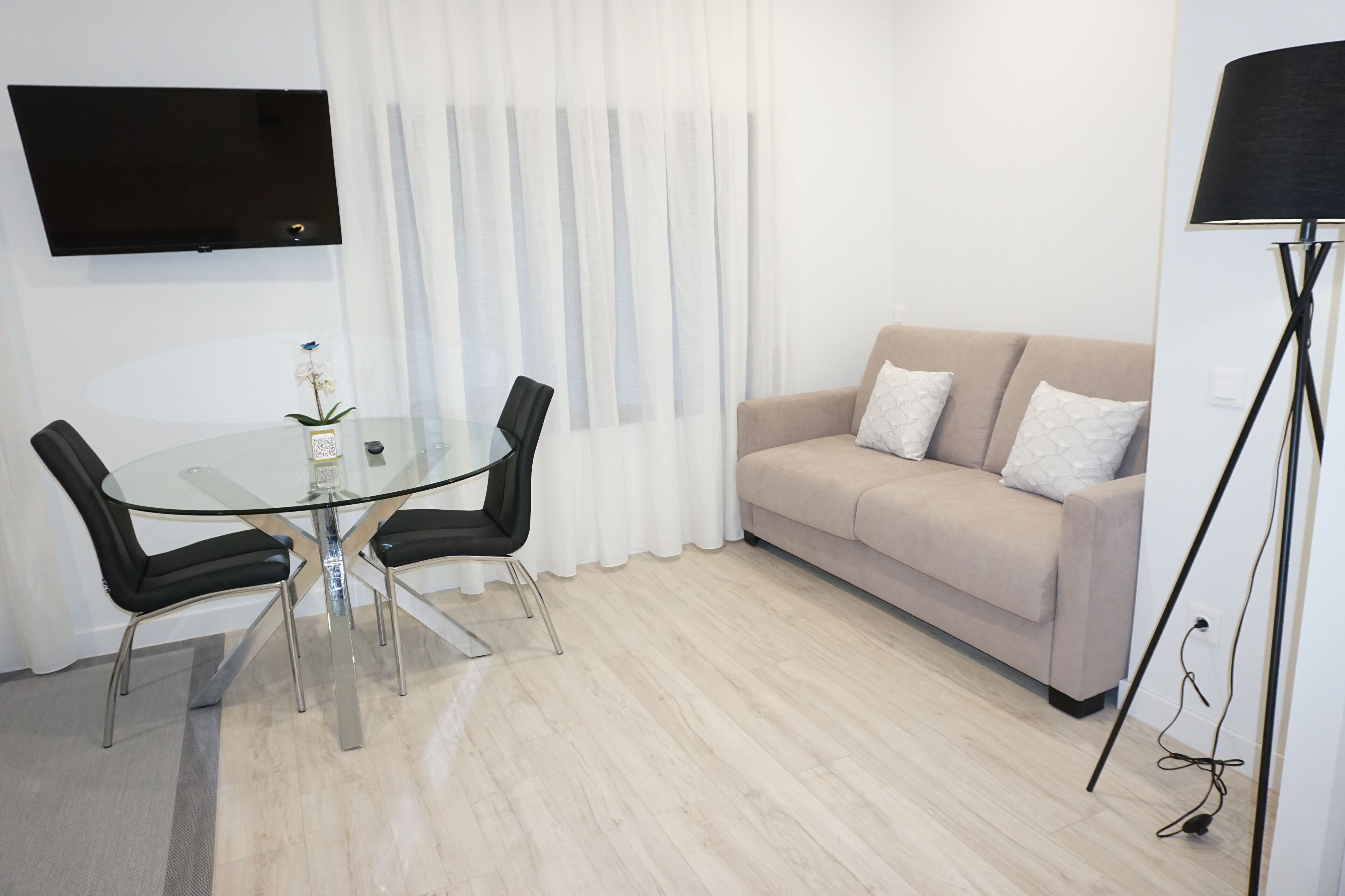Een moderne woonkamer met een glazen eettafel, twee zwarte stoelen, een beige bank met witte kussens, een flatscreen televisie aan de muur en een vloerlamp.