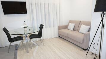 A modern living room with a glass dining table, two black chairs, a beige sofa with white pillows, a flat-screen TV mounted on the wall, and a floor lamp.