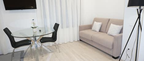 Een moderne woonkamer met een glazen eettafel, twee zwarte stoelen, een beige bank met witte kussens, een flatscreen televisie aan de muur en een vloerlamp.