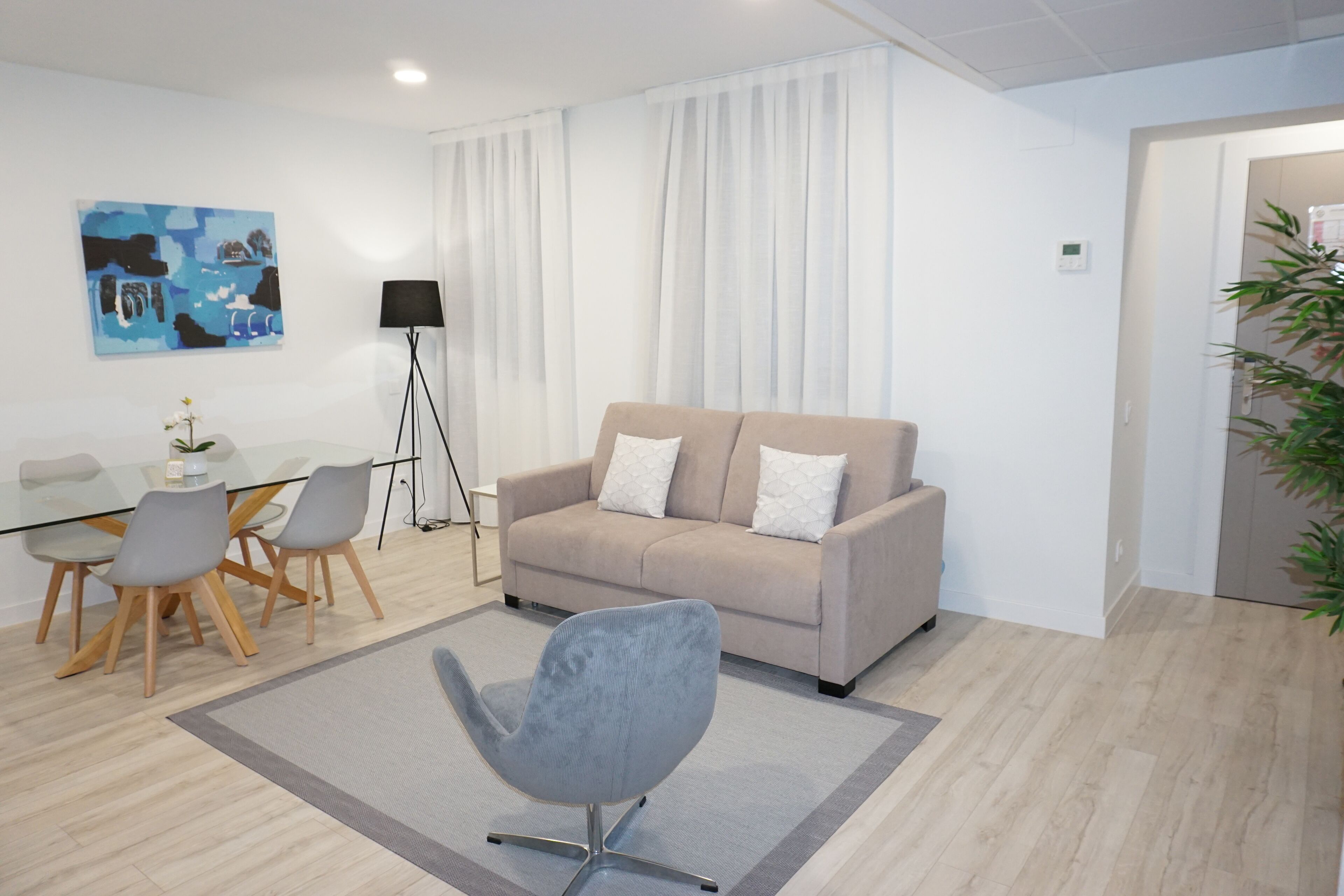A modern living room with a beige sofa, a glass dining table with chairs, a floor lamp, and a painting on the wall.
