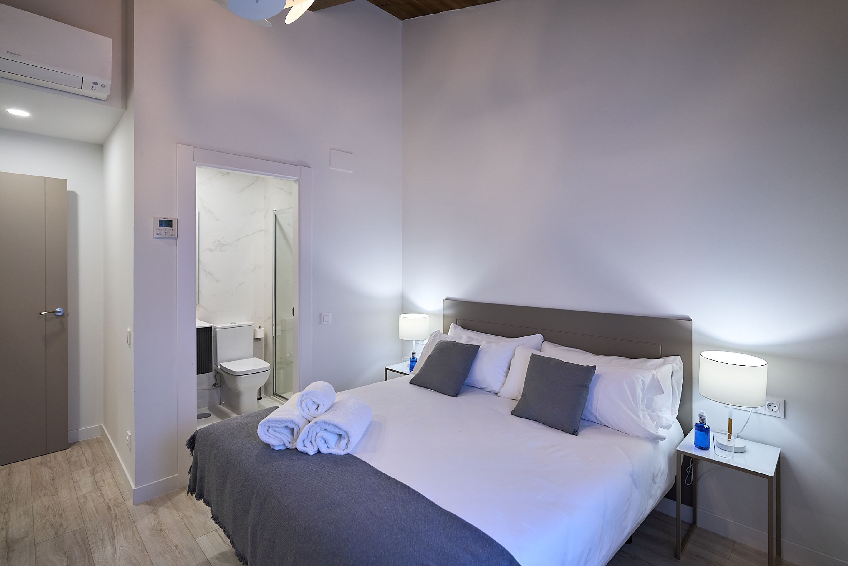 Une chambre d’hôtel comprenant un lit, une table de chevet avec une lampe, une salle de bain avec des toilettes et une douche, et une porte donnant sur une autre pièce.