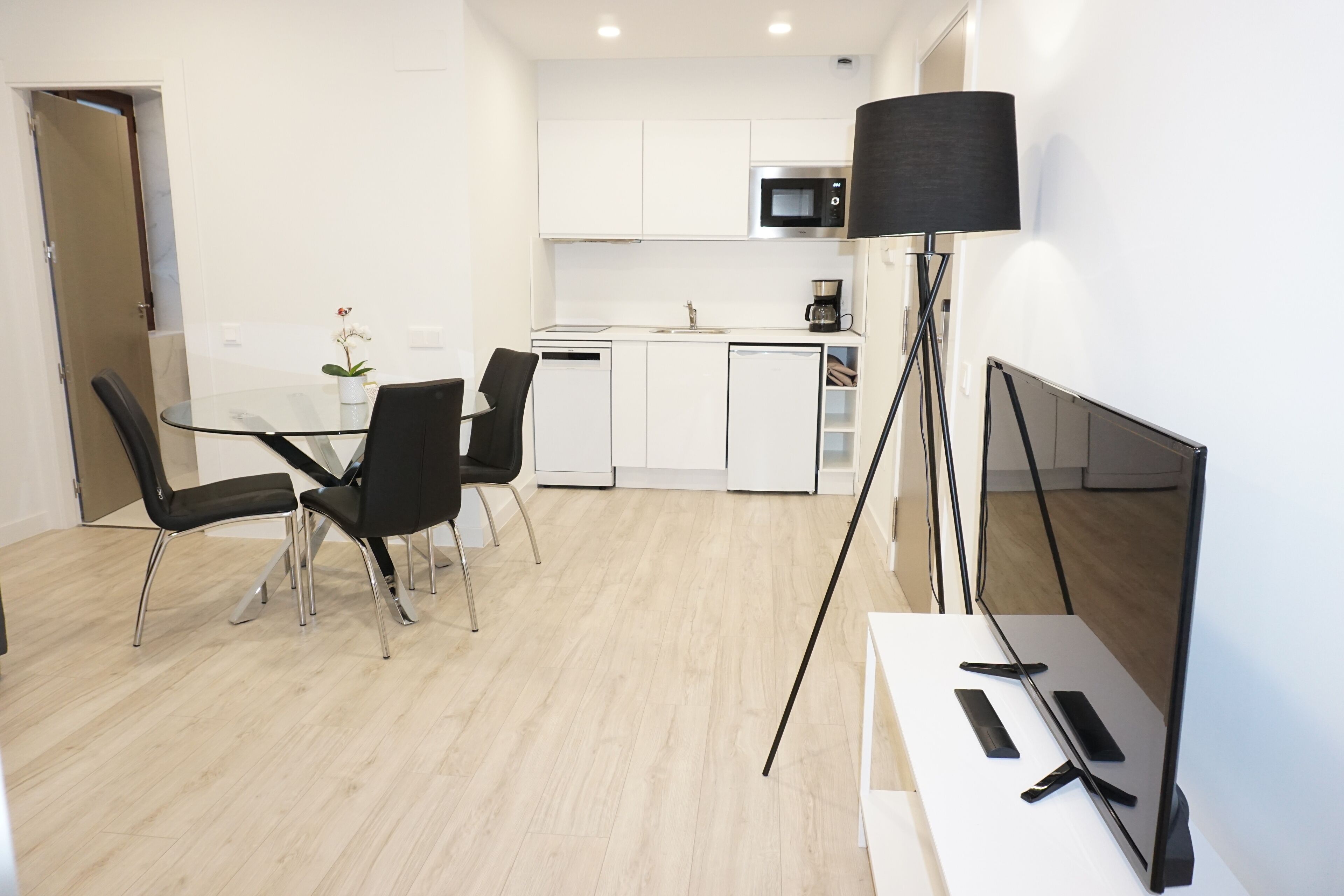 A modern kitchen with white cabinets, a microwave, and a coffee maker. There is a dining area with a glass table and black chairs. A floor lamp is visible in the corner.