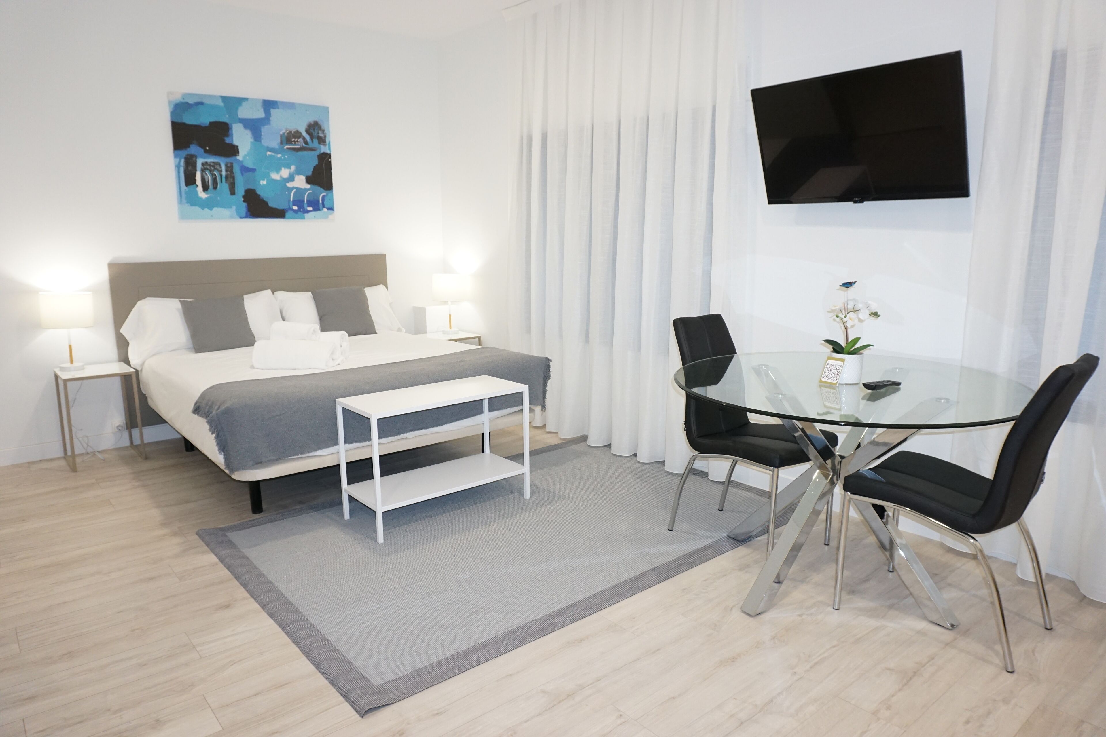 A modern hotel room with a bed, bedside tables, a glass table with chairs, a TV, and a painting on the wall.