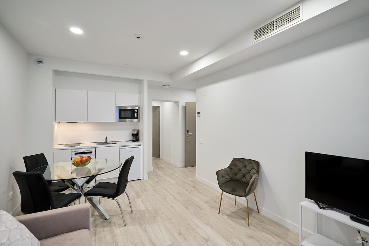 A modern living room with a glass dining table, black chairs, a grey sofa, a TV on a white stand, and a kitchen in the background.