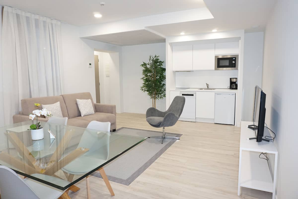 A modern living room with a glass dining table, a sofa, a chair, a potted plant, and a kitchen area with white cabinets and a microwave.