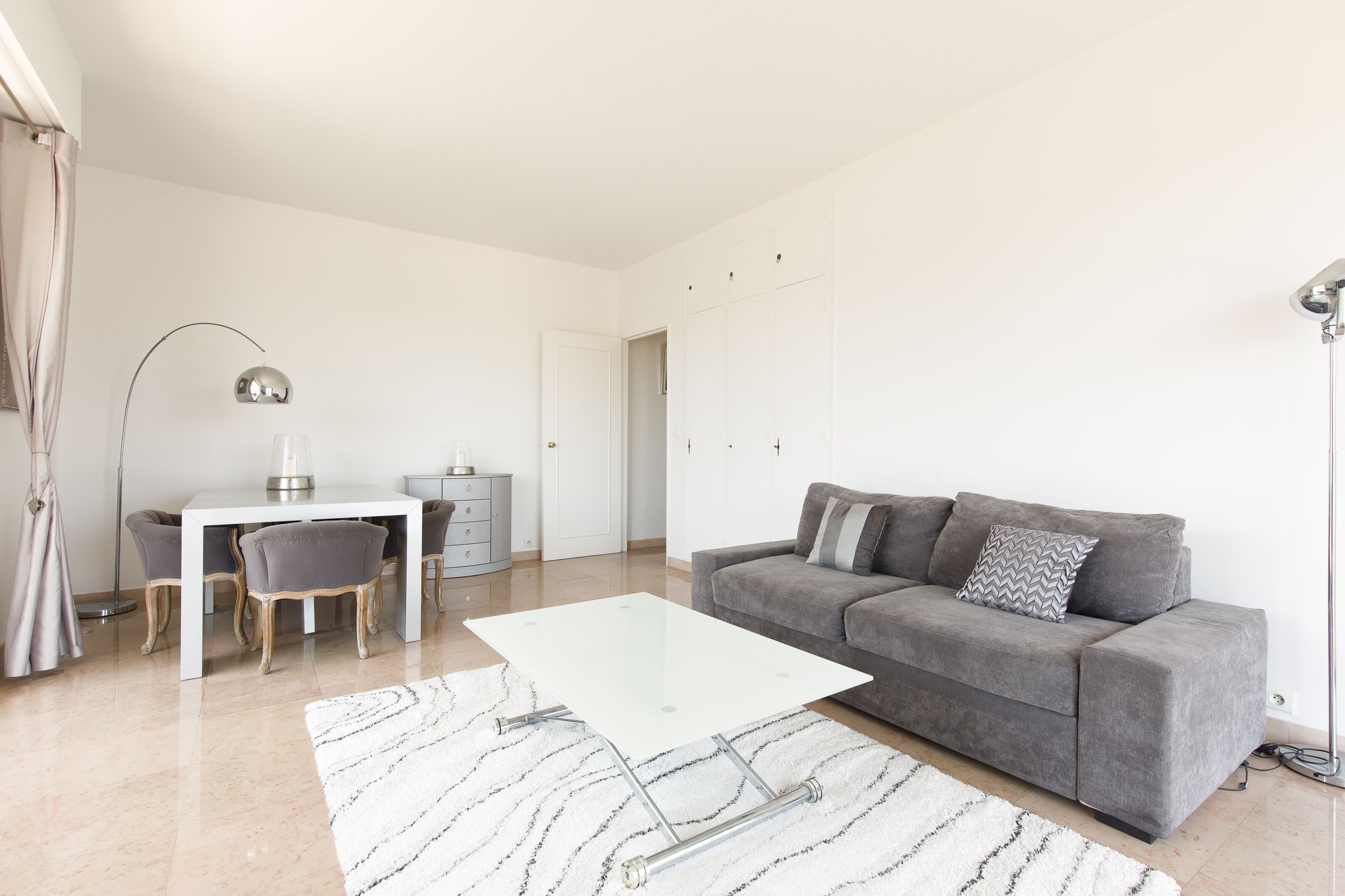 A modern living room with a grey sofa, a white coffee table, and a dining area with a white table and grey chairs.