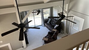 Een ruime woonkamer met een groot raam, een donkerbruine leren bank en twee plafondventilatoren.