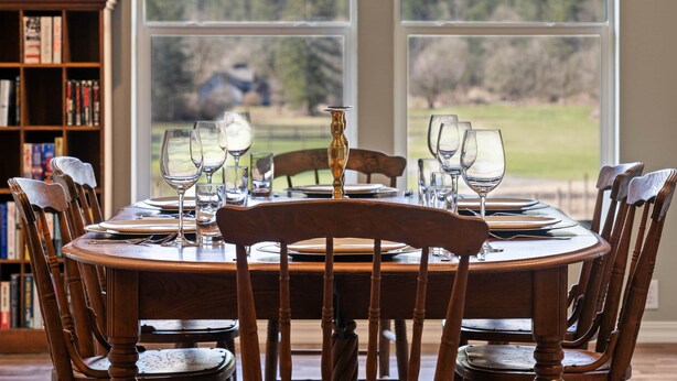 Mesa de comedor dispuesta para seis personas, con copas de vino y un candelabro, con vistas a un paisaje boscoso a través de amplias ventanas.
