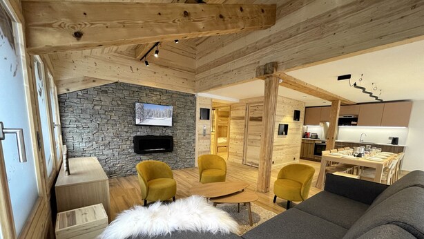Un accogliente soggiorno con parete in pietra, soffitto in legno e televisore. Sono presenti poltrone gialle, un divano grigio e un tavolo da pranzo in legno.