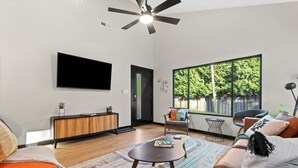 A modern living room with a flat-screen TV, a wooden cabinet, a sofa, a coffee table, and a large window with a view of trees. - Mid-Century 3 BR In Heart of Downtown Lex (Lexington)