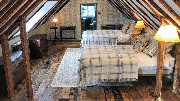 A bedroom with a sloped ceiling, wooden beams, a bed with a plaid blanket, two bedside lamps, a nightstand, a small table with a lamp, and a window. - 1 bedroom accommodation in Fernhurst (Fernhurst)