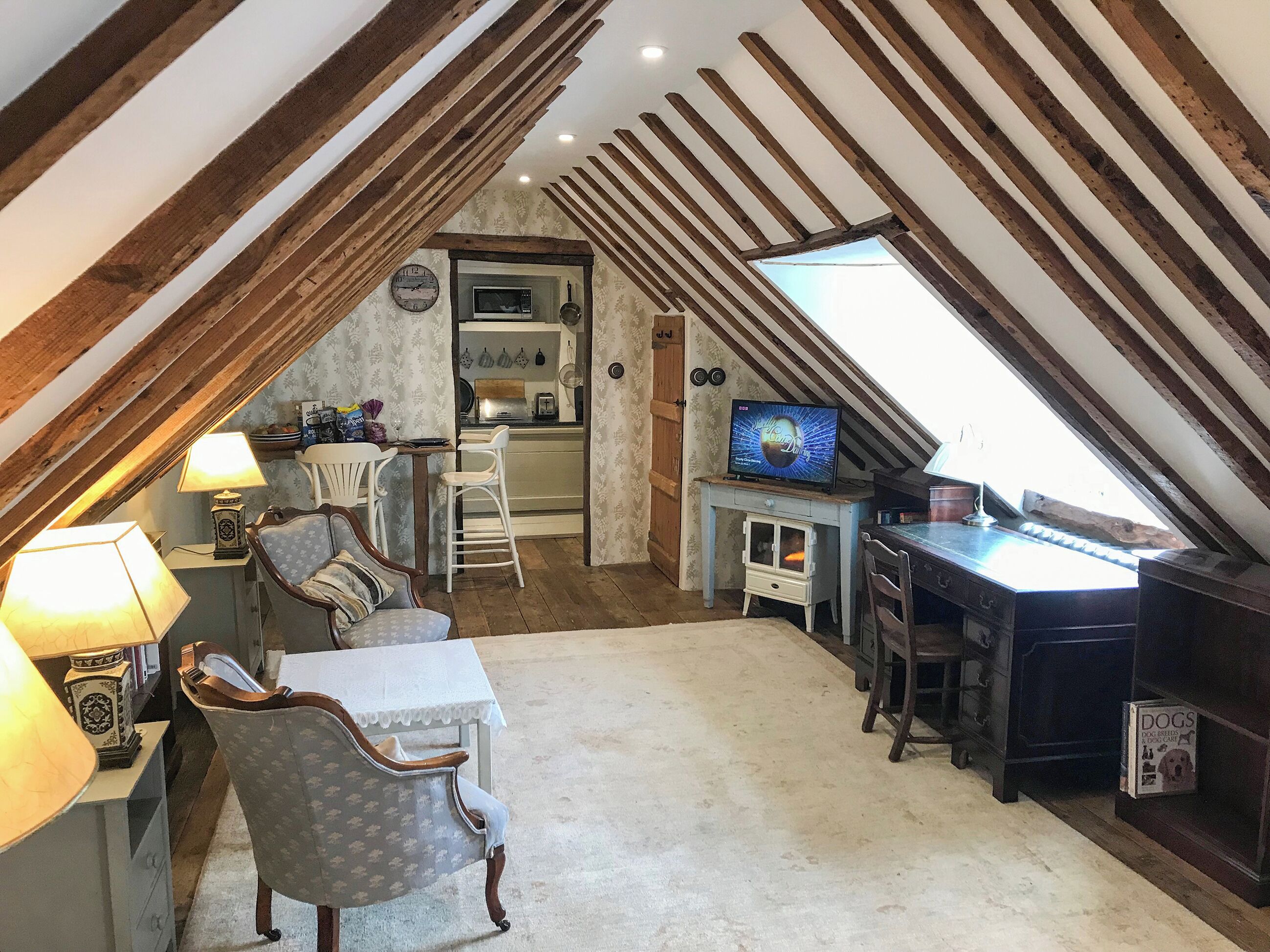 A cozy attic room with a sloped ceiling, a television, a dining table with chairs, and a small kitchenette.