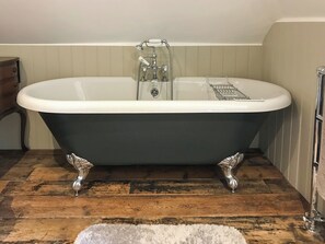 A clawfoot bathtub with a chrome faucet and shower head, set on a wooden floor with a white rug in front.